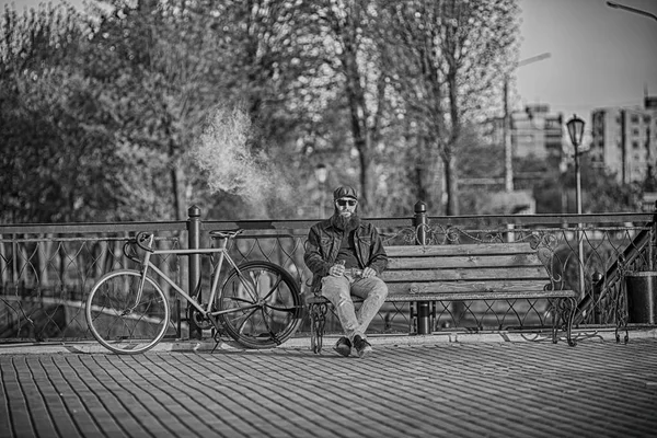 Vape na vida. Jovem com barba grande em óculos de sol e na tampa tendo um descanso e vaping um cigarro eletrônico perto de bicicleta correção vintage após passeio no banco no parque da cidade. Preto e branco . — Fotografia de Stock