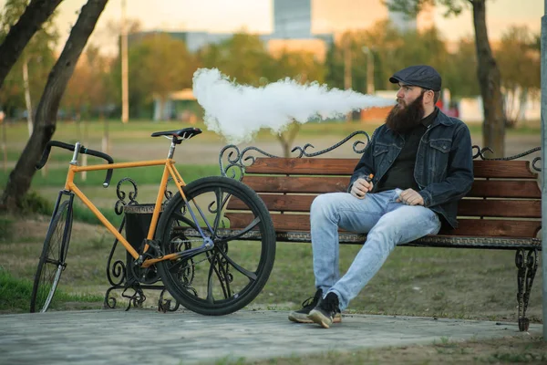 Vape na vida. Jovem com a barba grande gourgeous nos óculos de sol e na tampa que tem um descanso e vaping um cigarro eletrônico perto da bicicleta do reparo do vintage após o passeio no banco no parque da cidade . — Fotografia de Stock