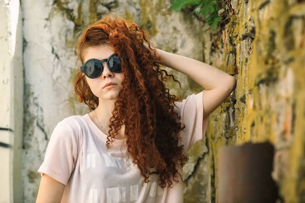 Hermosa adolescente. Retrato al aire libre de una hermosa joven blanca con el pelo rizado rojo en gafas de sol posando cerca de la antigua pared de ladrillo rojo destruido . —  Fotos de Stock