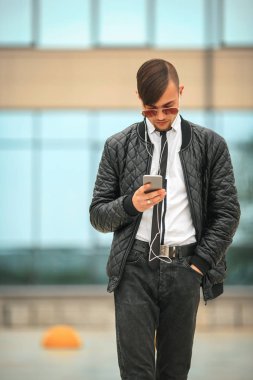 Portret havacı güneş gözlüğü içinde modern saç kesimi ile yakışıklı genç bir beyaz adam ve kulaklık ile şehir arka plan karşısında hareket eden telefon.