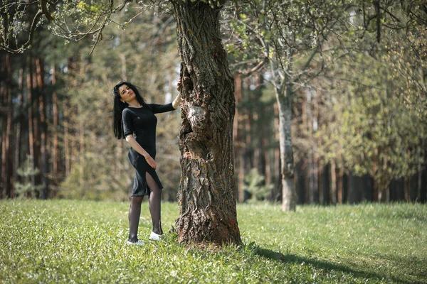 Портрет молодої красивої білої дівчини з чорним волоссям і в чорному одязі, що позує біля старого дерева . — стокове фото