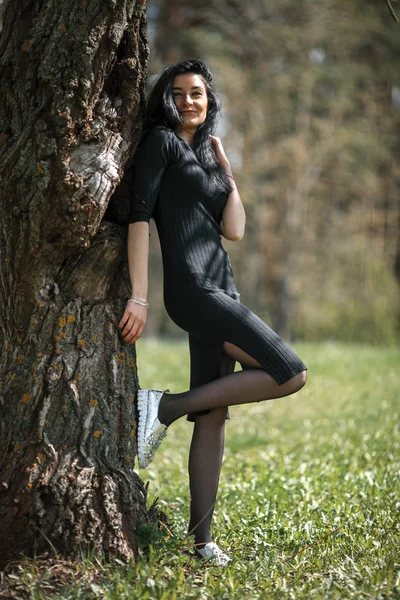 Retrato de chica blanca bonita joven con el pelo negro y en ropa negra posando cerca del árbol viejo . —  Fotos de Stock