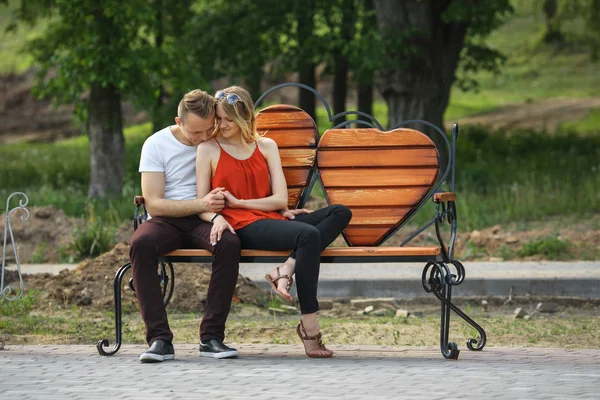 Pareja joven enamorándose en el banco en forma de corazones en el parque de la ciudad en el día de verano . —  Fotos de Stock