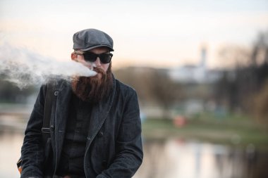 Vape adam gerçek hayatta sakallı. Şapka ve güneş gözlüğü vaping elektronik sigara büyük sakallı genç adam portresi puffs bahar akşam nehirde yakınındaki dolgu üzerinde buhar ile.