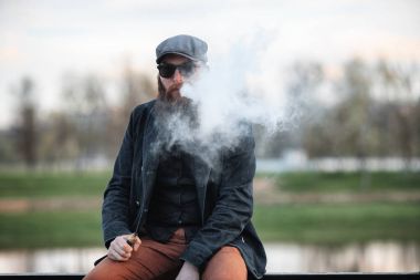 Vape adam gerçek hayatta sakallı. Şapka ve güneş gözlüğü vaping elektronik sigara büyük sakallı genç adam portresi puffs bahar akşam nehirde yakınındaki dolgu üzerinde buhar ile.