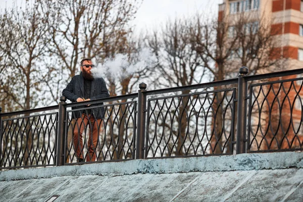 Vape adam gerçek hayatta sakallı. Güneş gözlüğü vaping elektronik sigara büyük sakallı genç adam portresi puffs yaz aylarında gün batımında dolgu üzerinde buhar ile.