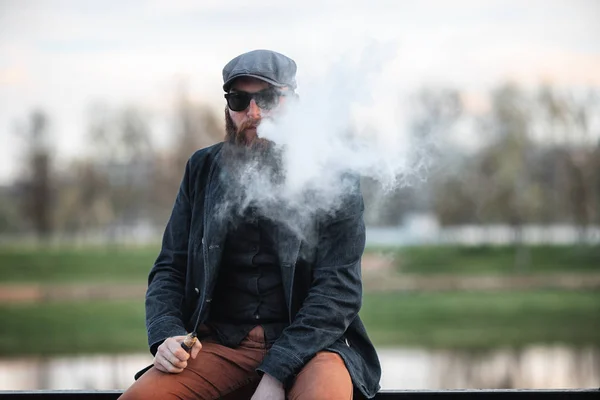 Vape barbudo homem na vida real. Retrato de jovem com barba grande em um boné e óculos de sol vaping um cigarro eletrônico com sopros de vapor no dique perto do rio na noite de primavera . — Fotografia de Stock