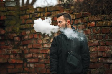 Vape adam. Fotoğraf siyah elbiseli vaping ve eski yakınındaki elektronik sigara buhar kapalı icar modern saç kesimi ile yakışıklı bir genç beyaz adamın kırmızı tuğla duvar yıkıldı. Yaşam tarzı.