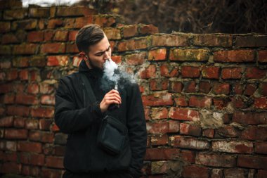 Vape adam. Fotoğraf siyah elbiseli vaping ve eski yakınındaki elektronik sigara buhar kapalı icar modern saç kesimi ile yakışıklı bir genç beyaz adamın kırmızı tuğla duvar yıkıldı. Yaşam tarzı.