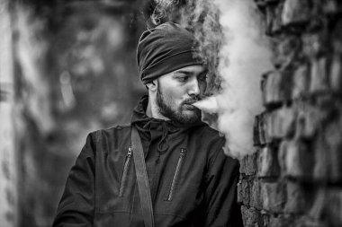 Vape adam. Fotoğraf şapka ve siyah elbiseli vaping ve eski yıkılan tuğla duvar yakınındaki elektronik sigara buhar kapalı icar yakışıklı genç bir beyaz adam. Siyah ve beyaz fotoğraf.