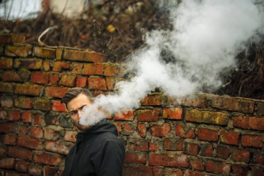 Vape adam. Fotoğraf siyah elbiseli vaping ve eski yakınındaki elektronik sigara buhar kapalı icar modern saç kesimi ile yakışıklı bir genç beyaz adamın kırmızı tuğla duvar yıkıldı. Yaşam tarzı.
