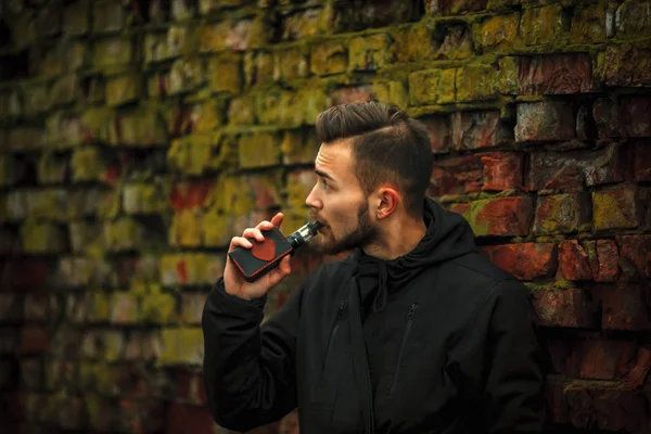 Vape adam. Fotoğraf siyah elbiseli vaping yakınındaki eski elektronik sigara içinde modern saç kesimi ile yakışıklı bir genç beyaz adamın kırmızı tuğla duvar yıkıldı. Yaşam tarzı.