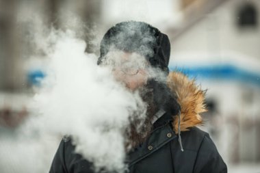 Vape adam gerçek hayatta sakallı. Büyük genç adam portresi sakal gözlük ve siyah kap vaping elektronik sigara ve Buhar icar içinde belgili tanımlık kış.