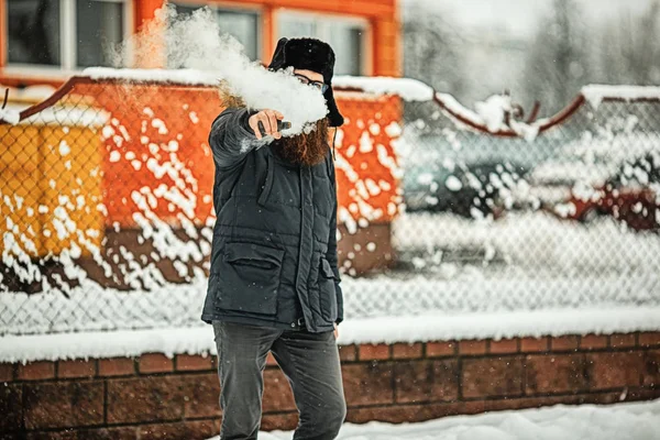 Vape bearded man in real life. Portrait of young guy with large beard in glasses and a cap vaping an electronic cigarette and letting out steam opposite the fence in the winter.