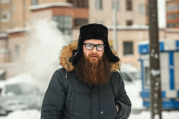 Vape bearded man in real life. Portrait of young guy with large beard in glasses and a black cap vaping an electronic cigarette and letting out steam in the winter.
