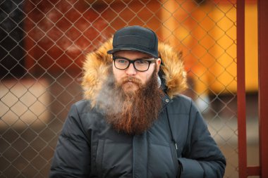 Vape adam gerçek hayatta sakallı. Gözlük ve bir kap vaping elektronik sigara ve Buhar çitin karşısında sonbaharda izin büyük sakallı genç adam portresi.