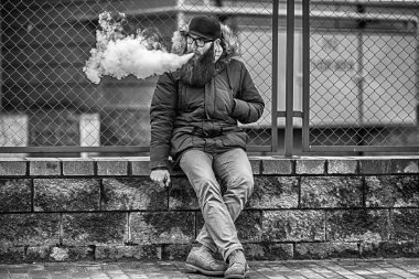 Vape adam gerçek hayatta sakallı. Genç portresi elektronik sigara içinde çit karşısında gözlük ve bir kap vaping büyük sakallı adam