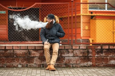 Vape adam gerçek hayatta sakallı. Gözlük ve bir kap vaping elektronik sigara ve Buhar çitin karşısında sonbaharda izin büyük sakallı genç adam portresi.
