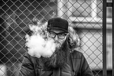 Vape adam gerçek hayatta sakallı. Gözlük ve bir kap vaping elektronik sigara sonbaharında çitin karşısında büyük sakallı genç adam portresi. Siyah ve beyaz.