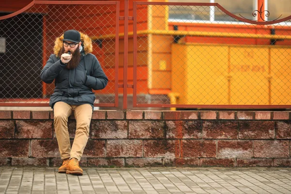 Vape bearded man in real life. Portrait of young guy with large beard in glasses and a cap vaping an electronic cigarette opposite the fence in the