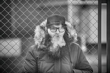 Vape adam gerçek hayatta sakallı. Gözlük ve bir kap vaping elektronik sigara sonbaharında çitin karşısında büyük sakallı genç adam portresi. Siyah ve beyaz.