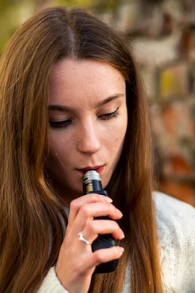Vaping teenager. Young pretty white caucasian teenage girl with problem skin smoking an electronic cigarette the ruined brick wall on the street in the autumn. Bad habit. Vape activity. Close up.