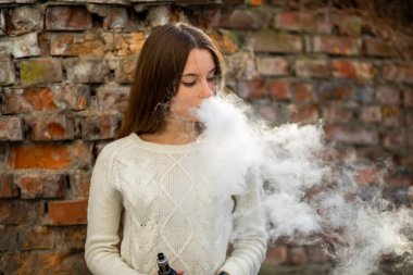Buharlı genç. Genç, beyaz, beyaz, sorunlu genç kız sonbaharda caddenin yıkılmış tuğla duvarının yanında elektronik sigara içiyor. Kötü alışkanlık. Vestiyer etkinliği.