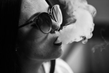 Buharlı genç. Güneş gözlüklü, beyaz tenli, evde sigara içen beyaz, genç bir kız. Kötü alışkanlık. Vape. Siyah ve beyaz. Kapatın. Siyah ve beyaz