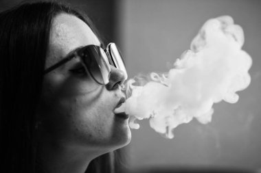 Buharlı genç. Güneş gözlüklü, beyaz tenli, evde sigara içen beyaz, genç bir kız. Kötü alışkanlık. Vape. Siyah ve beyaz. Kapatın. Siyah ve beyaz