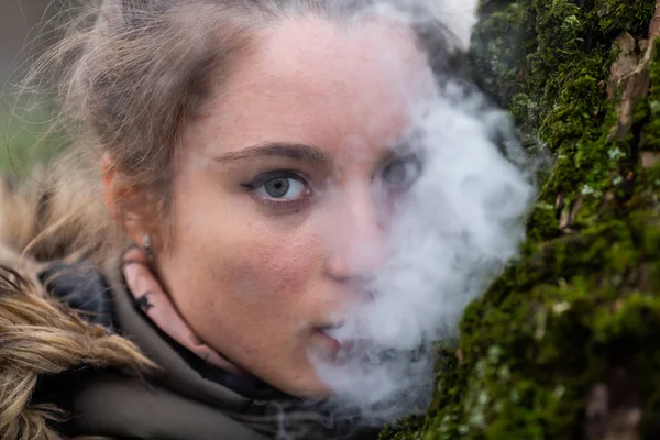 Vaping teenager. Young pretty white caucasian teenage girl in green jacket with hood smoking an electronic cigarette on the street in the park near the tree in the autumn day. Bad habit. Vape activity