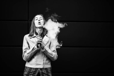 Buharlı genç. Gündelik giysiler içinde genç, beyaz bir kız sonbaharda karanlık ve modern bir grubun karşısında elektronik sigara içiyor. Kötü alışkanlık. Vestiyer aktivitesi. Siyah ve beyaz.