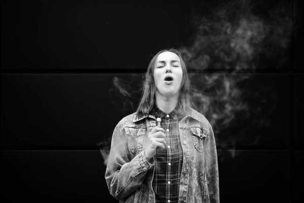 Vaping teenager. Young pretty white girl in casual clothes smoking an electronic cigarette opposite dark modern backgroun on the street in the autumn. Bad habit. Vape activity. Black and white.