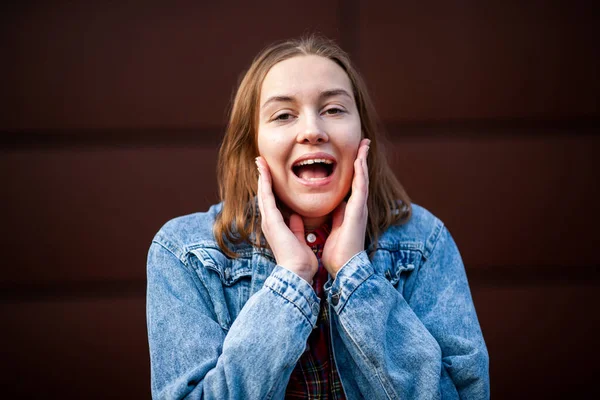 Emotions. Happy. Teenager. Young pretty white caucasian girl in casual jean jacket shows tongue opposite dark modern backgroun on the street in the autumn.