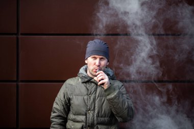 Vape adam. Güz aylarında modern kahverengi arka planda elektronik sigara içen sıradan giysili beyaz beyaz sakallı genç bir adam. Çok kötü bir alışkanlık..
