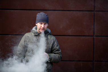 Vape adam. Güz aylarında modern kahverengi arka planda elektronik sigara içen sıradan giysili beyaz beyaz sakallı genç bir adam. Çok kötü bir alışkanlık..