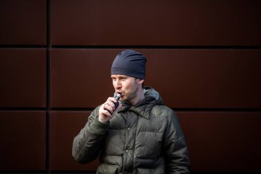 Vape adam. Güz aylarında modern kahverengi arka planda elektronik sigara içen sıradan giysili, yakışıklı, sakallı beyaz bir adam. Çok kötü bir alışkanlık..