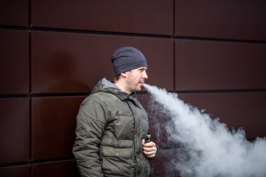 Vape adam. Güz aylarında modern kahverengi arka planda elektronik sigara içen sıradan giysili beyaz beyaz sakallı genç bir adam. Çok kötü bir alışkanlık..