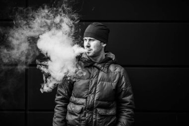 Vape adam. Güz aylarında modern arka planda elektronik sigara içen sıradan giysili beyaz beyaz sakallı genç bir adam. Çok kötü bir alışkanlık. Siyah ve beyaz.