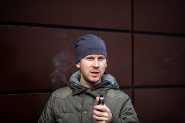 Vape adam. Güz aylarında modern kahverengi arka planda elektronik sigara içen sıradan giysili, yakışıklı, sakallı beyaz bir adam. Çok kötü bir alışkanlık..