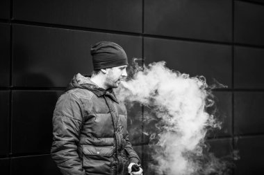 Vape adam. Güz aylarında modern arka planda elektronik sigara içen sıradan giysili beyaz beyaz sakallı genç bir adam. Çok kötü bir alışkanlık. Siyah ve beyaz.