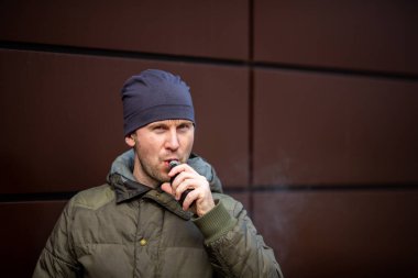 Vape adam. Güz aylarında modern kahverengi arka planda elektronik sigara içen sıradan giysili, yakışıklı, sakallı beyaz bir adam. Çok kötü bir alışkanlık..