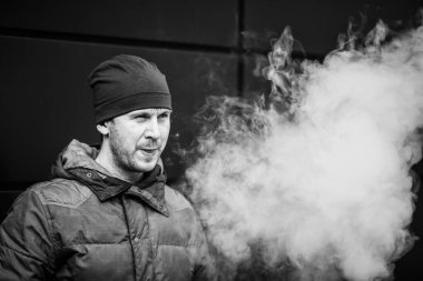 Vape adam. Güz aylarında modern arka planda elektronik sigara içen sıradan giysili beyaz beyaz sakallı genç bir adam. Çok kötü bir alışkanlık. Siyah ve beyaz.