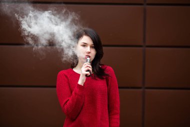 Vape teenager. Young pretty white caucasian girl in a red sweater smoking an electronic cigarette opposite modern brown background on the street in the winter. Deadly bad habit.