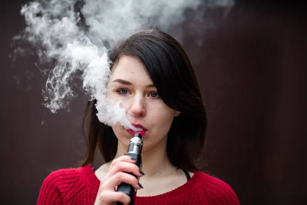 Vape teenager. Young pretty white caucasian girl in a red sweater smoking an electronic cigarette opposite modern brown background on the street in the winter. Deadly bad habit.
