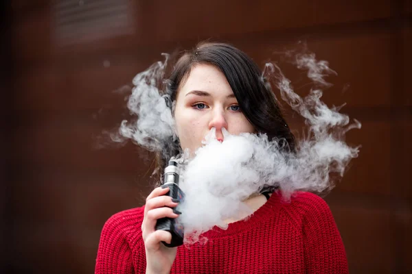 Vape teenager. Young pretty white caucasian girl in a red sweater smoking an electronic cigarette opposite modern brown background on the street in the winter. Deadly bad habit.