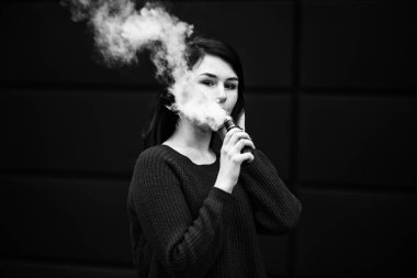 Vape teenager. Young pretty white caucasian girl in a sweater smoking an electronic cigarette opposite modern brown background on the street in the winter. Deadly bad habit. Black and white