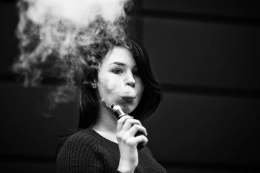 Derisi sorunlu bir genci buharlaştırmak. Ceketli, beyaz, genç ve güzel bir kız akşamları sokakta modern kahverengi arka planda elektronik sigara içiyor. Çok kötü bir alışkanlık. Siyah ve beyaz