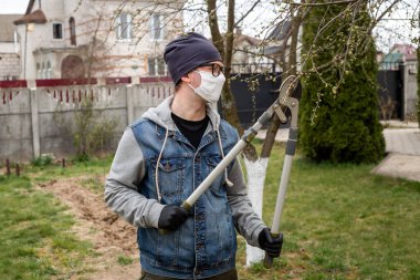 Bahçıvanlık. Beyaz maskeli, gözlüklü ve siyah eldivenli bir adam arka bahçesindeki ağaçların dallarını kesiyor. COVID-19. Avrupa 'da Coronavirus. Salgın yaşam.