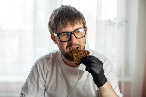 Govd パンデミックの間 台所で彼の手の中にパンのスライスとメガネ 白いマスクと黒の手袋の若い白人男性 ヨーロッパのコロナウイルス — ストック写真
