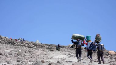 three male porters carrying heavy loads and supplies to the base camp at the foot of the summit of Mount Kilimanjaro clipart